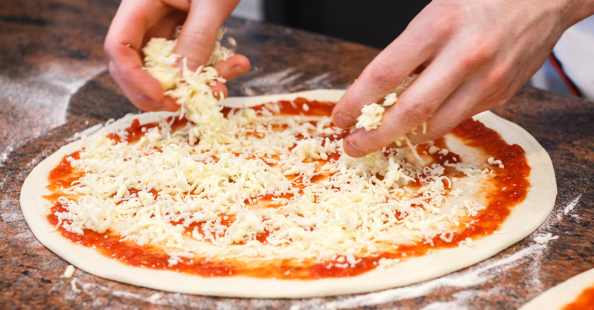 Hands adding a topping to a pizza.