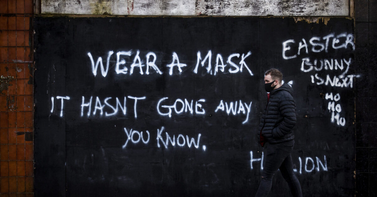 A man wearing a face covering walks past graffiti on the Lower Newtownards Road in Belfast with a message reading 'Wear a mask, it hasn't gone away you know' Friday, Jan. 1, 2021. (Liam McBurney/PA via AP)