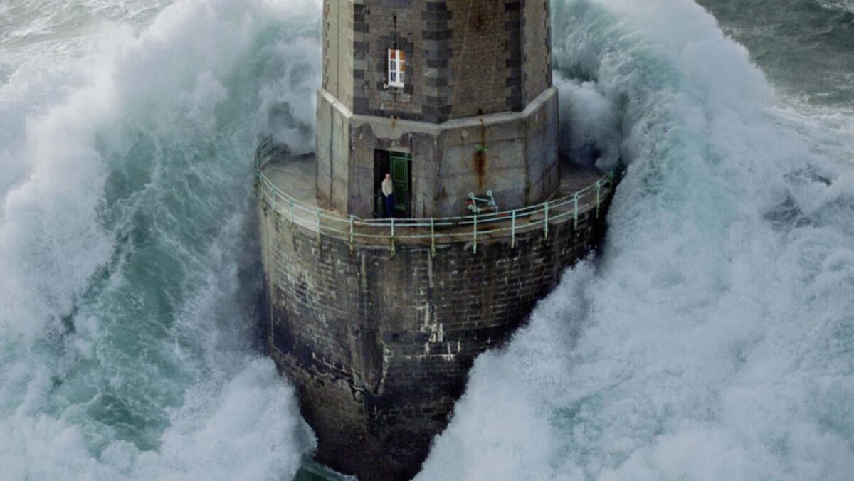 A photograph shows a big wave hitting a lighthouse while a lighthouse keeper stands in the doorway and the picture is from 1989 at the La Jument Lighthouse in France.