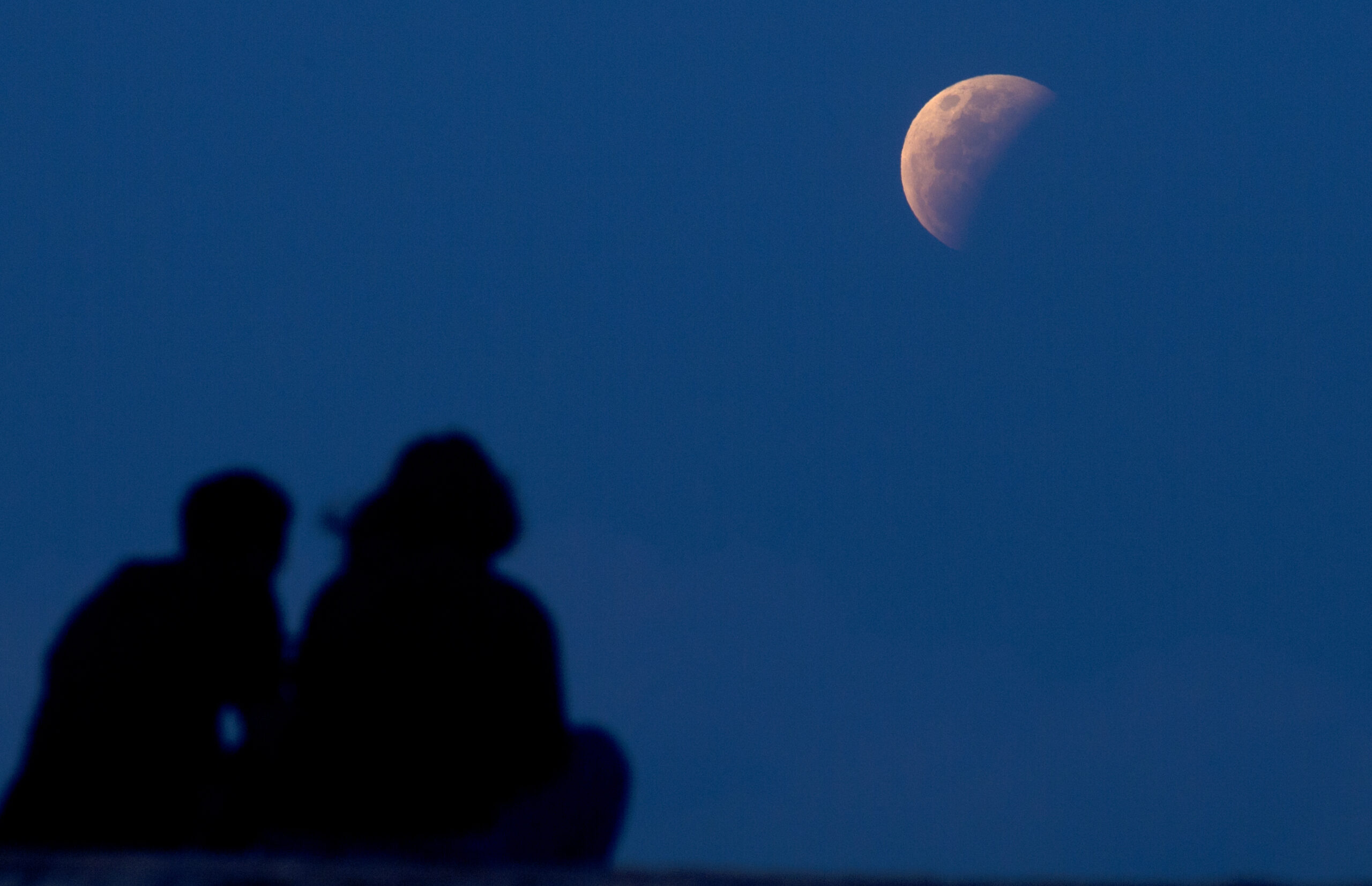 A couple watch the lunar eclipse at Sanur beach in Bali, Indonesia on Wednesday, May 26, 2021. The total lunar eclipse, also known as a super blood moon, is the first in two years with the reddish-orange color the result of all the sunrises and sunsets in Earth's atmosphere projected onto the surface of the eclipsed moon. (AP Photo/Firdia Lisnawati)