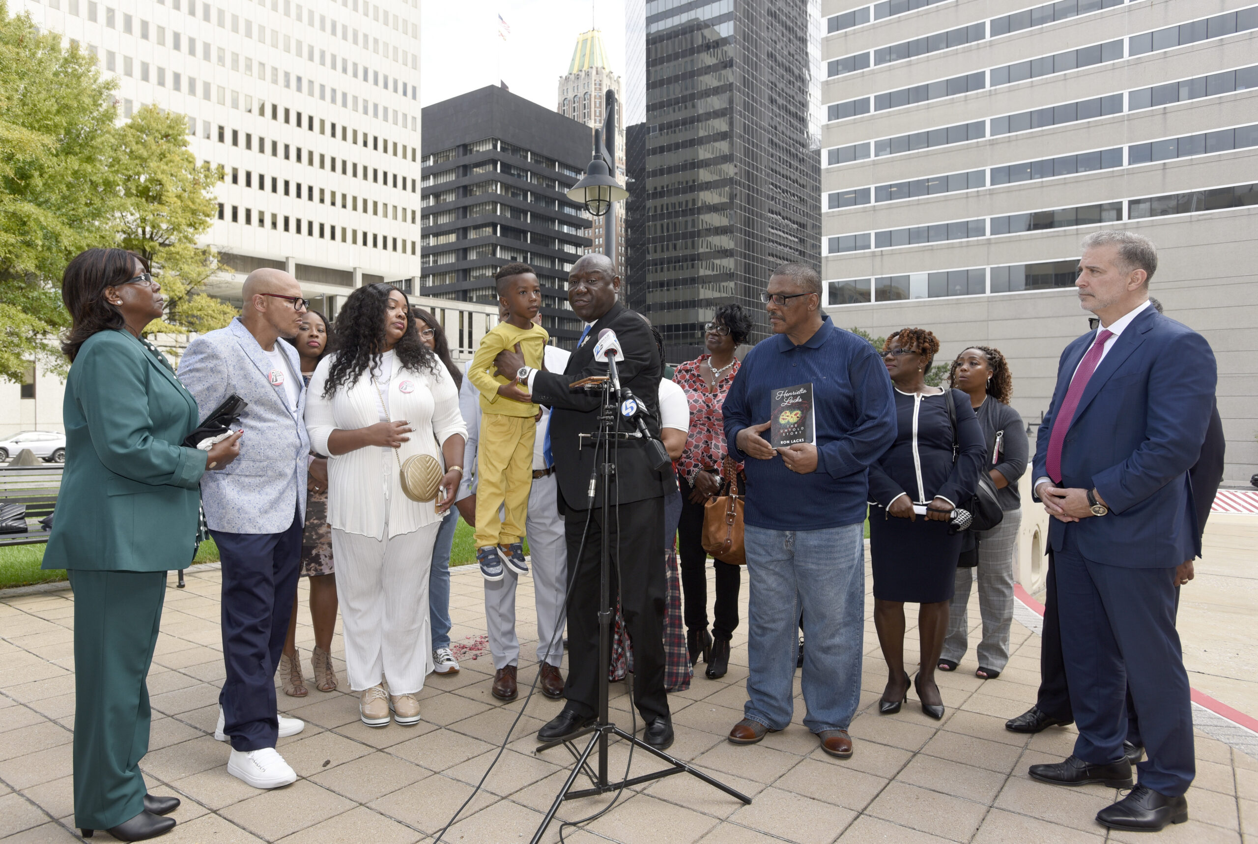 Attorney Ben Crump, center, holds Zayden Joseph, 6, the great-grandson of Henrietta Lacks, while standing with attorneys and other descendants of Lacks, whose cells have been used in medical research without her permission, outside the federal courthouse in Baltimore, Monday, Oct. 4, 2021. They announced during a news conference that Lacks' estate is filing a lawsuit against Thermo Fisher Scientific for using Lacks' cells, known as HeLa cells. (AP Photo/Steve Ruark)