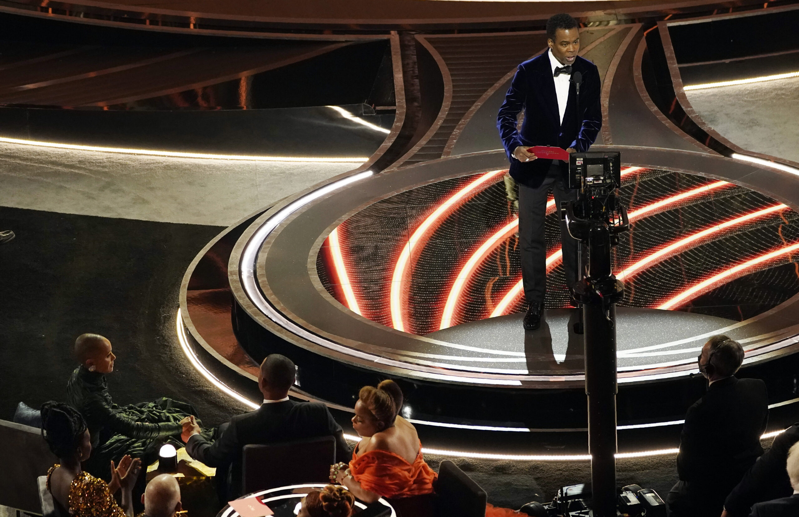 Presenter Chris Rock, right, speaks onstage as Jada Pinkett Smith and Will Smith, bottom left, look on after Smith went onstage and slapped Rock at the Oscars, Sunday, March 27, 2022, at the Dolby Theatre in Los Angeles. (AP Photo/Chris Pizzello)