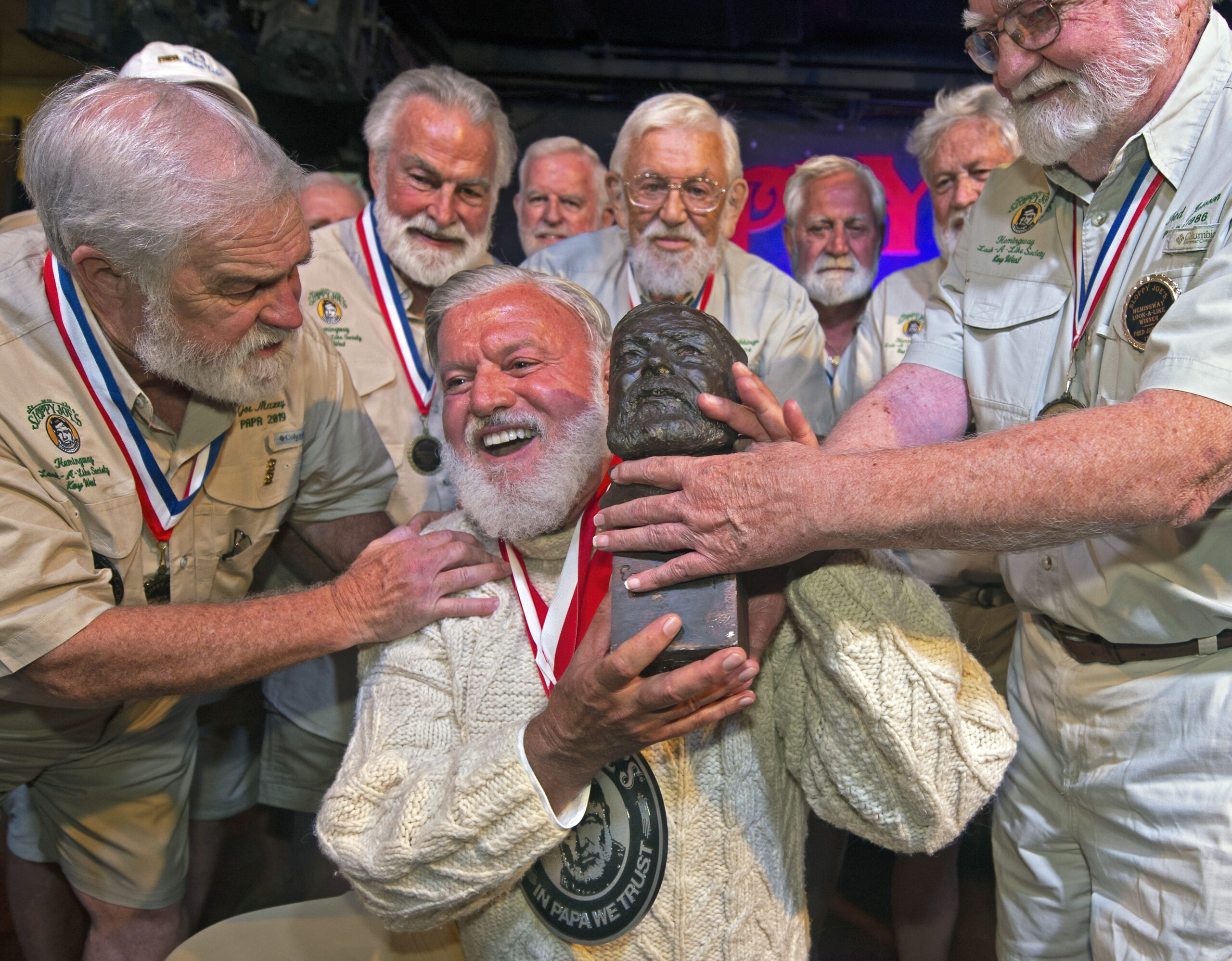 In this Saturday, July 23, 2022, photo provided by the Florida Keys News Bureau, Jon Auvil, center, receives an Ernest Hemingway bust and congratulations after he won the 2022 Hemingway Look-Alike Contest at Sloppy Joe's Bar in Key West, Fla. Left of Auvil is Joe Maxey, the 2019 winner, and at right is Fred Johnson, who won in 1986. Auvil, who finally won on his eighth attempt, beat 124 other entrants in the contest that was the highlight event of Key West's annual Hemingway Days festival that ends Sunday, July 24. Hemingway lived and wrote in Key West during most of the 1930s. (Andy Newman/Florida Keys News Bureau via AP)
