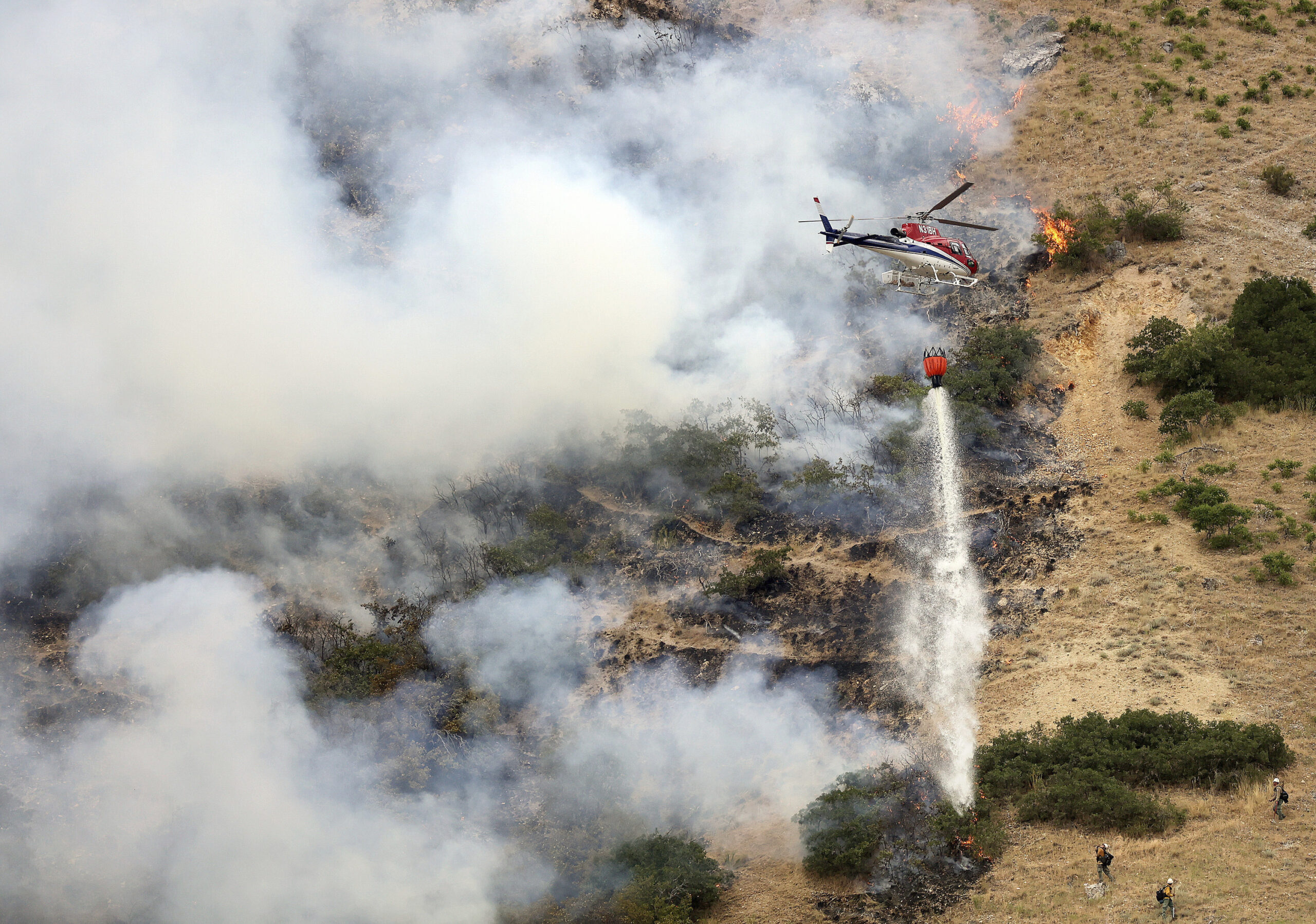 Firefighters battle a wildfire from the ground as a helicopter drops water above them in Springville on Monday, Aug. 1, 2022. The fire started when a man tried to burn a spider with a lighter. (Kristin Murphy/The Deseret News via AP)